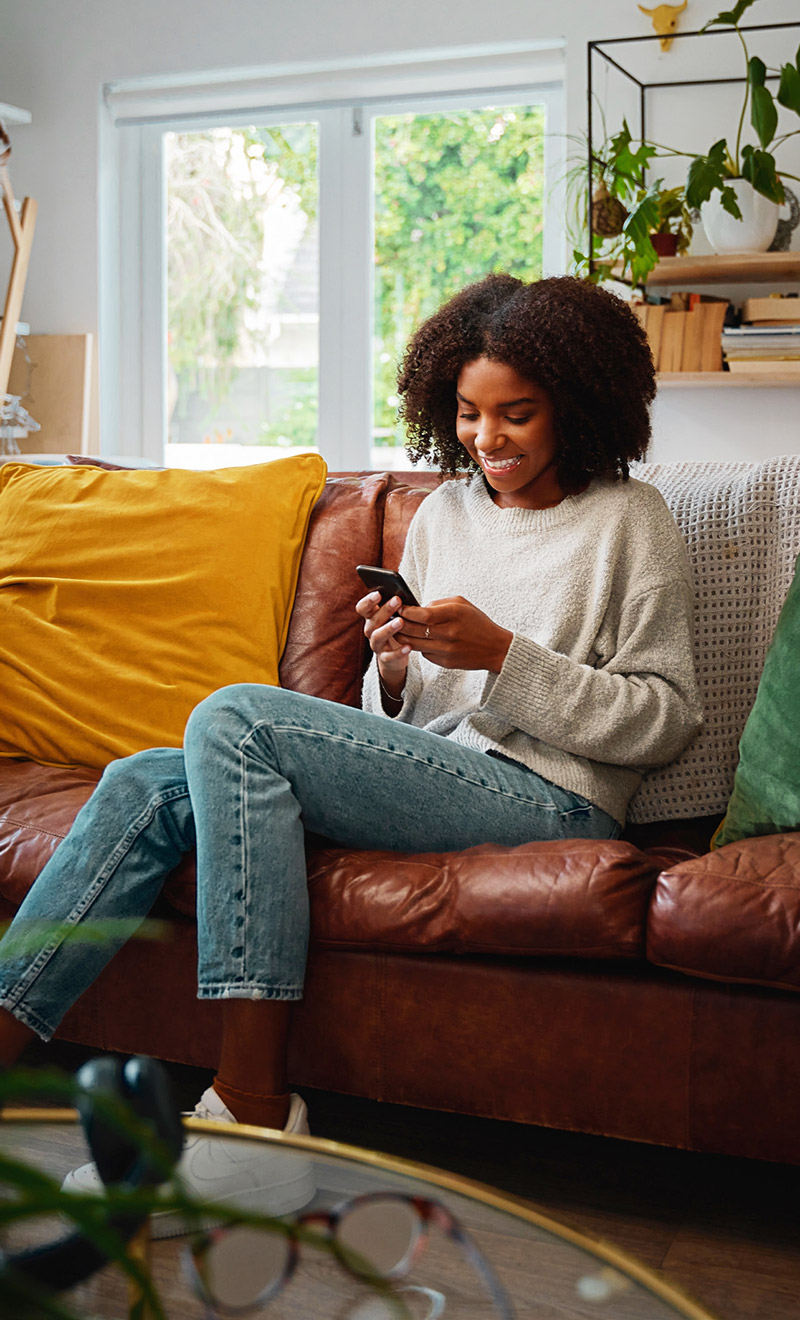 Young, black woman sitting on a couch smiling as she uses her mobile device Young, black woman sitting on a couch smiling as she uses her mobile device
